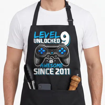 Cute level video game apron - Game controller with Level 9 Unlocked Awesome Since 2011 text and lock icon — shown on a black cotton canvas apron against a white background
