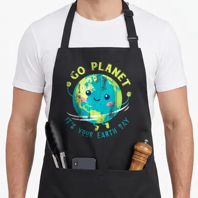Funny earth apron - Smiling cartoon earth with GO PLANET text and white orbit ring — shown on a black cotton canvas apron against a white background