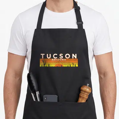 Arizona tucson apron - Retro Tucson Arizona text above orange yellow sunset stripes with grey cactus silhouettes — shown on a black cotton canvas apron against a white background