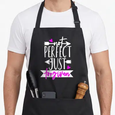 Arrow christian apron - Stacked text not perfect just forgiven with white arrows and two pink hearts — shown on a black cotton canvas apron against a white background