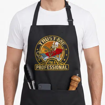 Wile coyote professional aprons - Wile E. Coyote on red rocket with Trust Me I'm A Professional text in orange circle — shown on a black cotton canvas apron against a white background