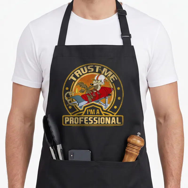 Wile coyote professional aprons - Wile E. Coyote on red rocket with Trust Me I'm A Professional text in orange circle — shown on a black cotton canvas apron against a white background