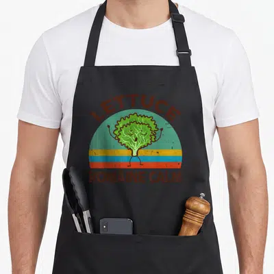 Lettuce vegan apron - Cartoon green lettuce character with arms raised under arched brown text and colorful horizontal — shown on a black cotton canvas apron against a white background