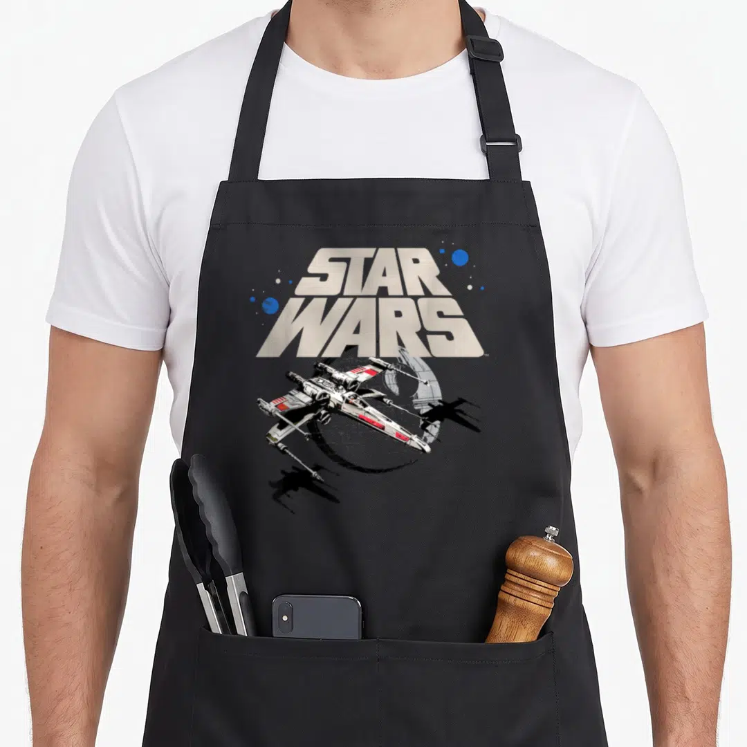 Approach x apron - Realistic X-wing starfighter below stacked STAR WARS text with blue planets and Death Star arc — shown on a black cotton canvas apron against a white background