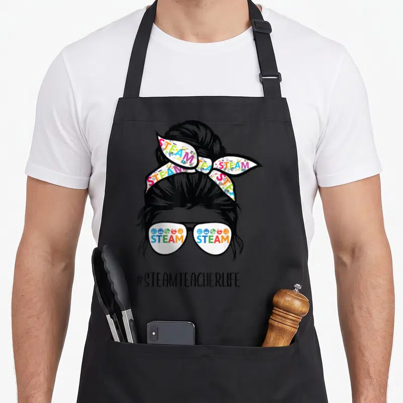 Steam messy aprons - Woman silhouette with STEAM text in glasses and headband, #STEAMTEACHERLIFE hashtag below — shown on a black cotton canvas apron against a white background