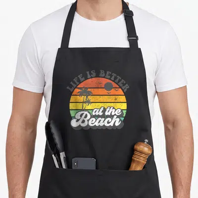 Better summer aprons - Retro sunset with palm trees and LIFE IS BETTER at the Beach text in orange yellow green and grey — shown on a black cotton canvas apron against a white background