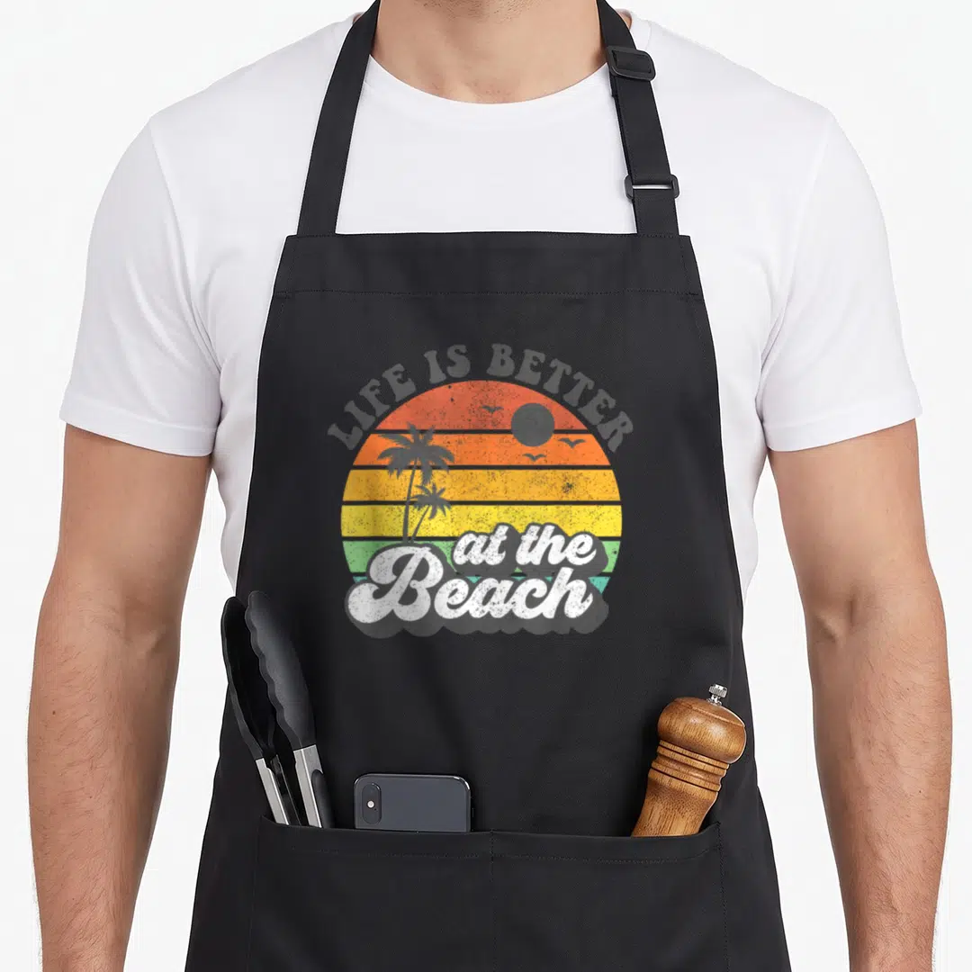 Better summer aprons - Retro sunset with palm trees and LIFE IS BETTER at the Beach text in orange yellow green and grey — shown on a black cotton canvas apron against a white background