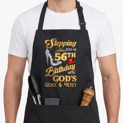 Step grace apron - Gold 56th birthday text with silver heel, crown, and red lips on black apron — shown on a black cotton canvas apron against a white background