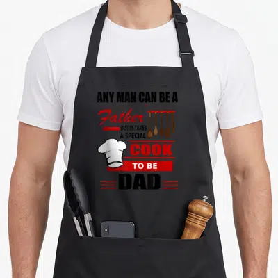 Father special aprons - Stacked text Any Man Can Be A Father with four brown cooking utensils and white chef hat — shown on a black cotton canvas apron against a white background