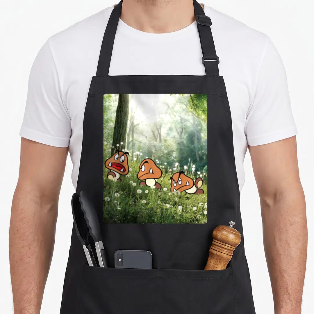 Goomba forest apron - Three cartoon mushroom characters with brown caps and white eyes hiding in green grass with white — shown on a black cotton canvas apron against a white background