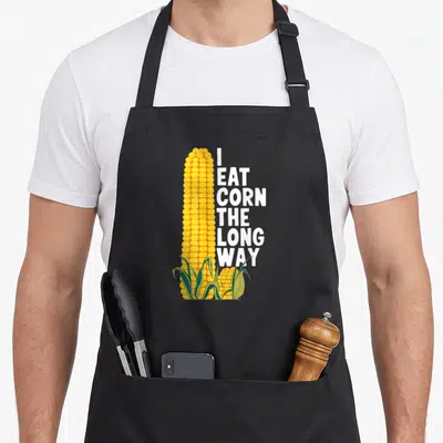 Inappropriate gag aprons - Vertical yellow corn ear with green leaves and stacked white text reading I eat corn — shown on a black cotton canvas apron against a white background