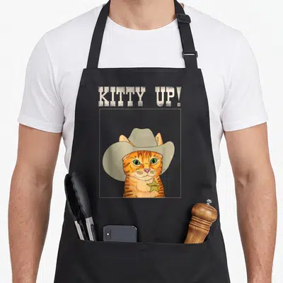 Cowboy kitty cat apron - Cartoon orange cat in cowboy hat and yellow sheriff badge under text KITTY UP! — shown on a black cotton canvas apron against a white background