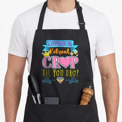 Retreat scrapbooking apron - SCRAPBOOKING Retreat CROP TIL YOU DROP text with crown, heart, diamond, and swirls — shown on a black cotton canvas apron against a white background