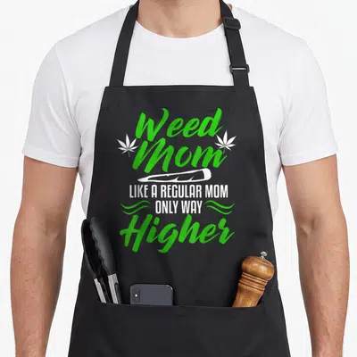 Weed golf mom apron - Green Weed Mom text with white leaves and joint graphic plus slogan LIKE A REGULAR MOM ONLY WAY — shown on a black cotton canvas apron against a white background