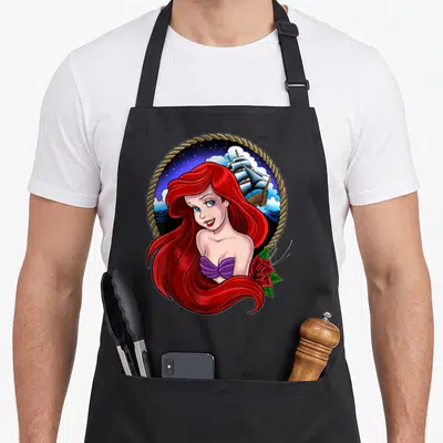 Ariel tattoo apron - Cartoon Ariel with red hair, purple top, red rose, ship, stars, rope border — shown on a black cotton canvas apron against a white background