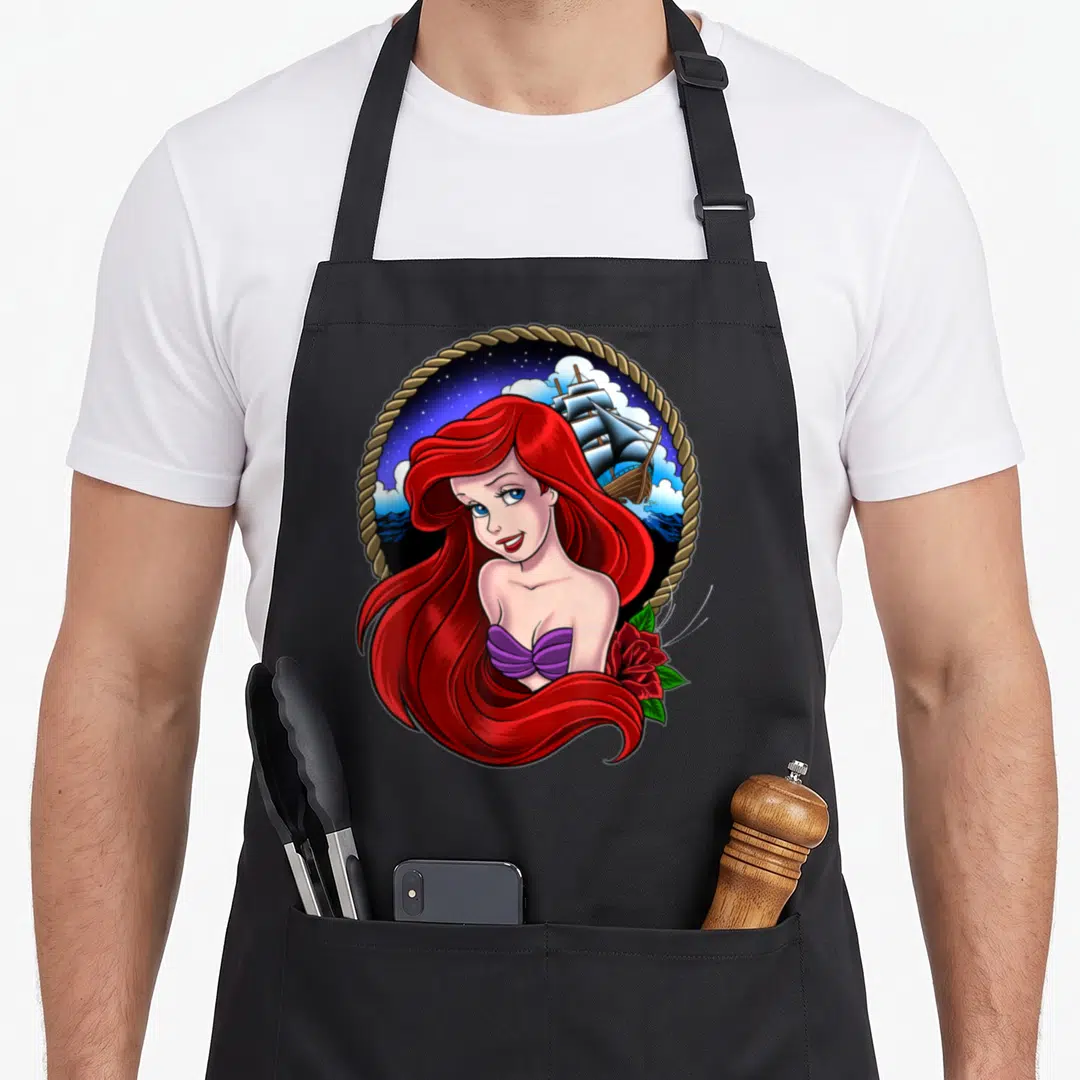 Ariel tattoo apron - Cartoon Ariel with red hair, purple top, red rose, ship, stars, rope border — shown on a black cotton canvas apron against a white background