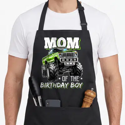 Funny birthday truck apron - Green monster truck graphic with MOM and OF THE BIRTHDAY BOY text — shown on a black cotton canvas apron against a white background