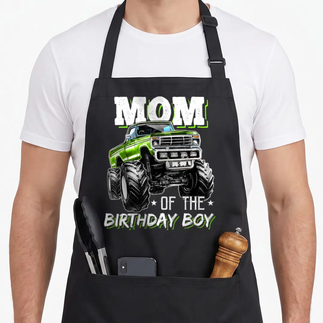 Funny birthday truck apron - Green monster truck graphic with MOM and OF THE BIRTHDAY BOY text — shown on a black cotton canvas apron against a white background