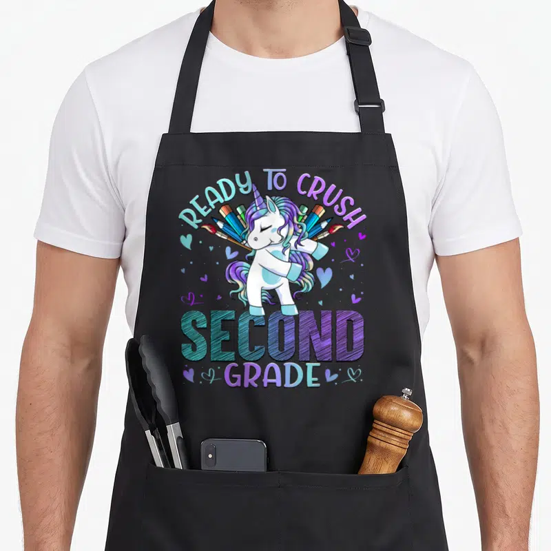 Ready crush 2nd apron - Cartoon unicorn with crossed pencils paintbrushes hearts and arched READY TO CRUSH SECOND GRADE — shown on a black cotton canvas apron against a white background