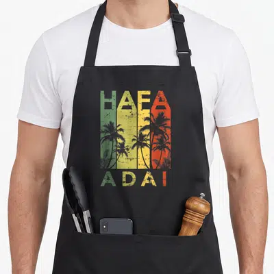 Funny hafa guam aprons - Retro HAFA ADAI text over green yellow red grey stripes with grey palm tree silhouettes — shown on a black cotton canvas apron against a white background