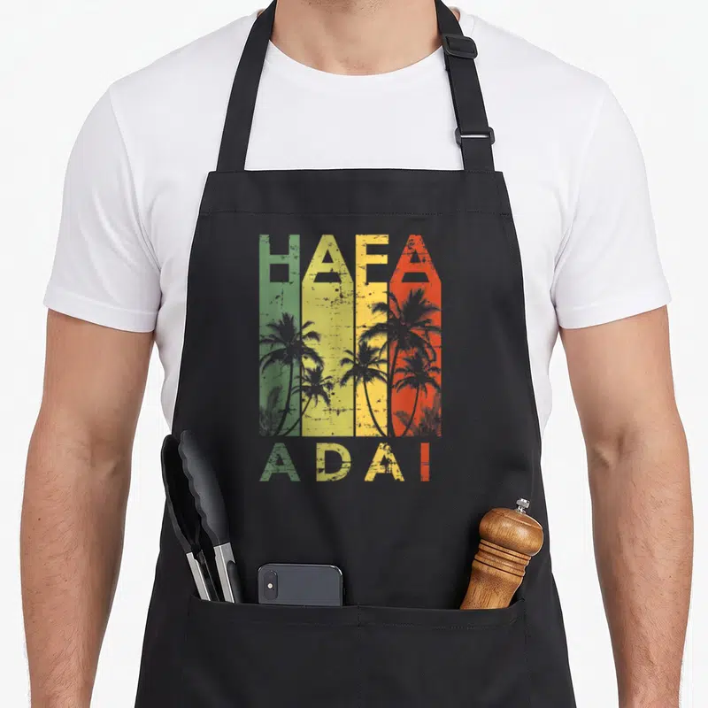Funny hafa guam aprons - Retro HAFA ADAI text over green yellow red grey stripes with grey palm tree silhouettes — shown on a black cotton canvas apron against a white background
