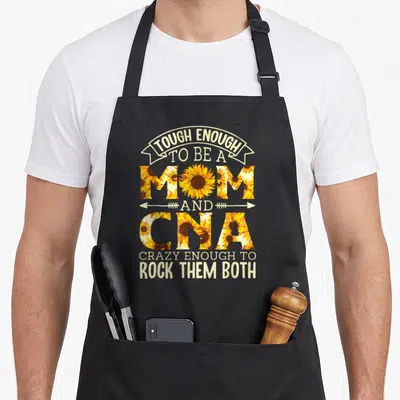 Tough mom apron - Text reading TOUGH ENOUGH TO BE A MOM AND CNA CRAZY ENOUGH TO ROCK THEM BOTH filled with sunflowers — shown on a black cotton canvas apron against a white background