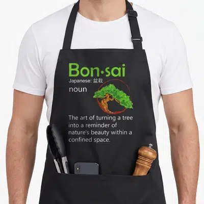 Definition tree apron - Green Bon·sai text with bonsai tree inside red circle and definition below — shown on a black cotton canvas apron against a white background