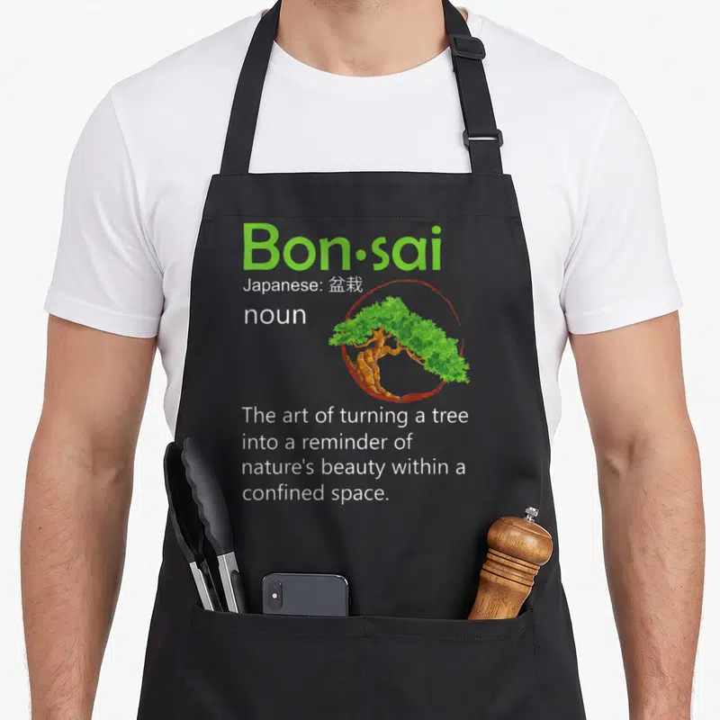 Definition tree apron - Green Bon·sai text with bonsai tree inside red circle and definition below — shown on a black cotton canvas apron against a white background