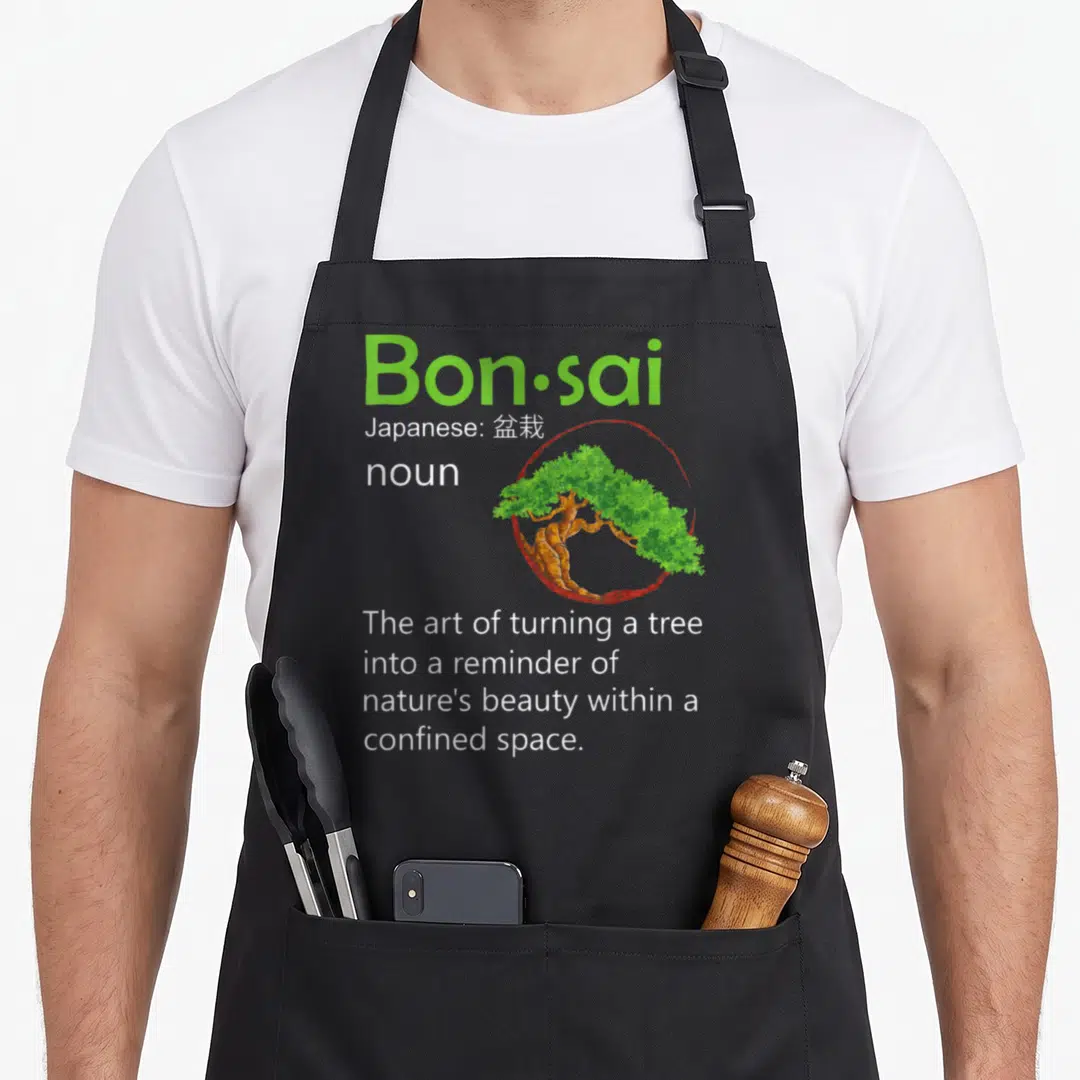 Definition tree apron - Green Bon·sai text with bonsai tree inside red circle and definition below — shown on a black cotton canvas apron against a white background