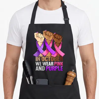 Breast domestic aprons - Three raised fists in different skin tones wrapped in pink and purple ribbons above stacked — shown on a black cotton canvas apron against a white background