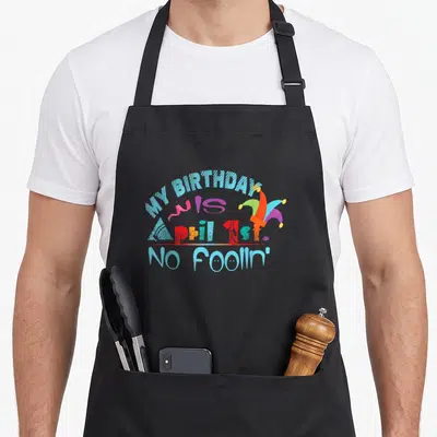 Fool birthday apron - Colorful jester hat and party horn next to arched text reading My Birthday is April 1st No Foolin — shown on a black cotton canvas apron against a white background