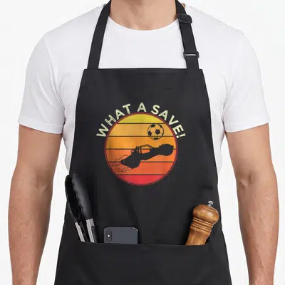 Rescue car apron - Retro sunset circle with soccer ball, rocket car silhouette, and text WHAT A SAVE — shown on a black cotton canvas apron against a white background