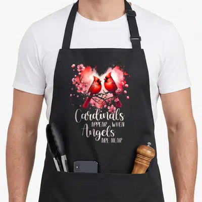 Cardinal bird apron - Two red cardinals on a branch inside a pink watercolor heart with stacked white text below — shown on a black cotton canvas apron against a white background