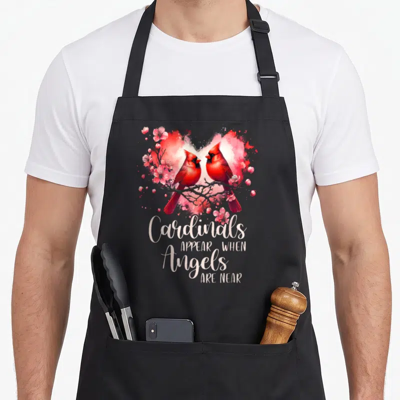 Cardinal bird apron - Two red cardinals on a branch inside a pink watercolor heart with stacked white text below — shown on a black cotton canvas apron against a white background