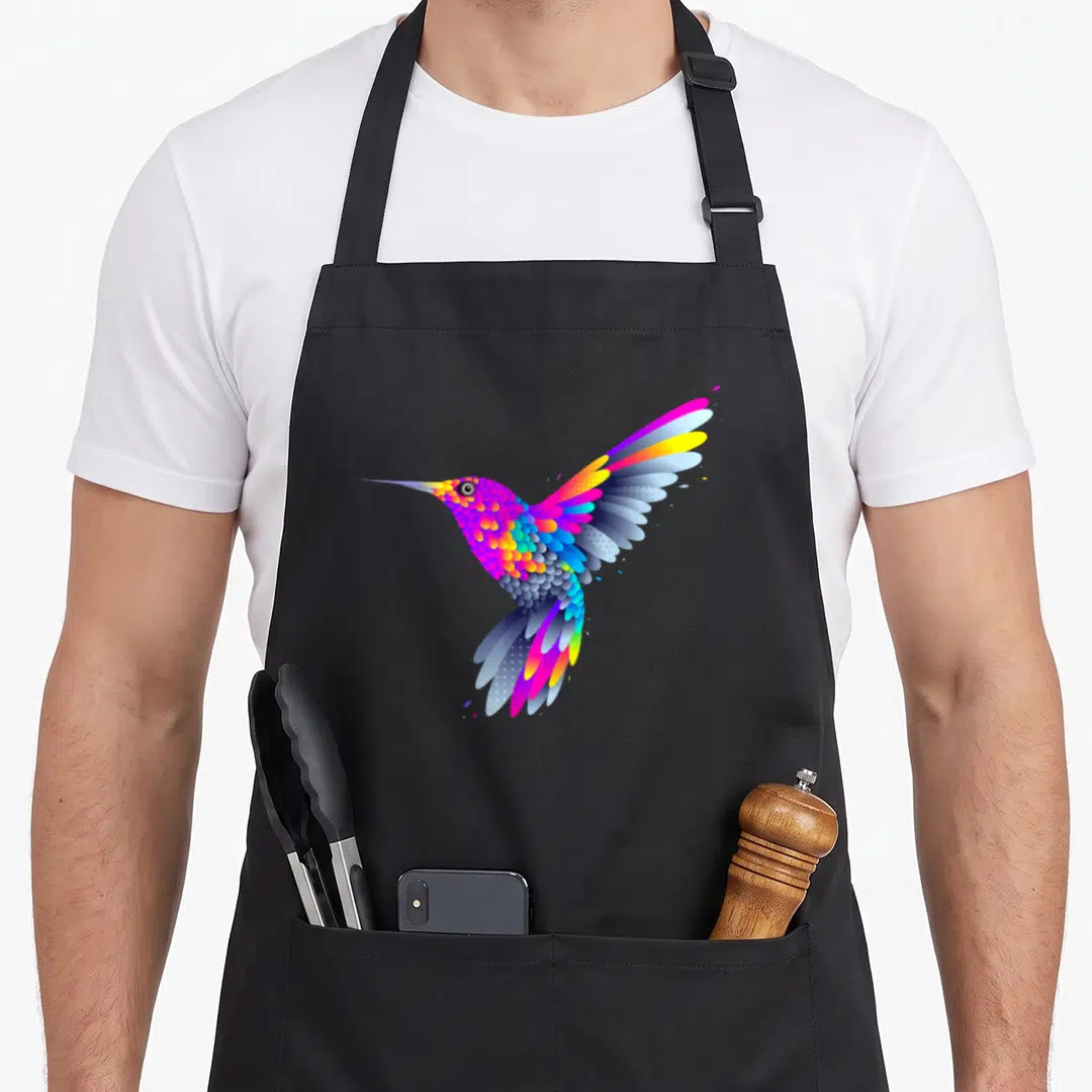 Peaceful colorful aprons - Stylized hummingbird in purple, blue, pink, and yellow cartoon feathers centered on black — shown on a black cotton canvas apron against a white background