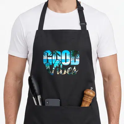 Positive beach apron - GOOD Vibes text filled with blue ocean, green palms, and tan sand photography — shown on a black cotton canvas apron against a white background