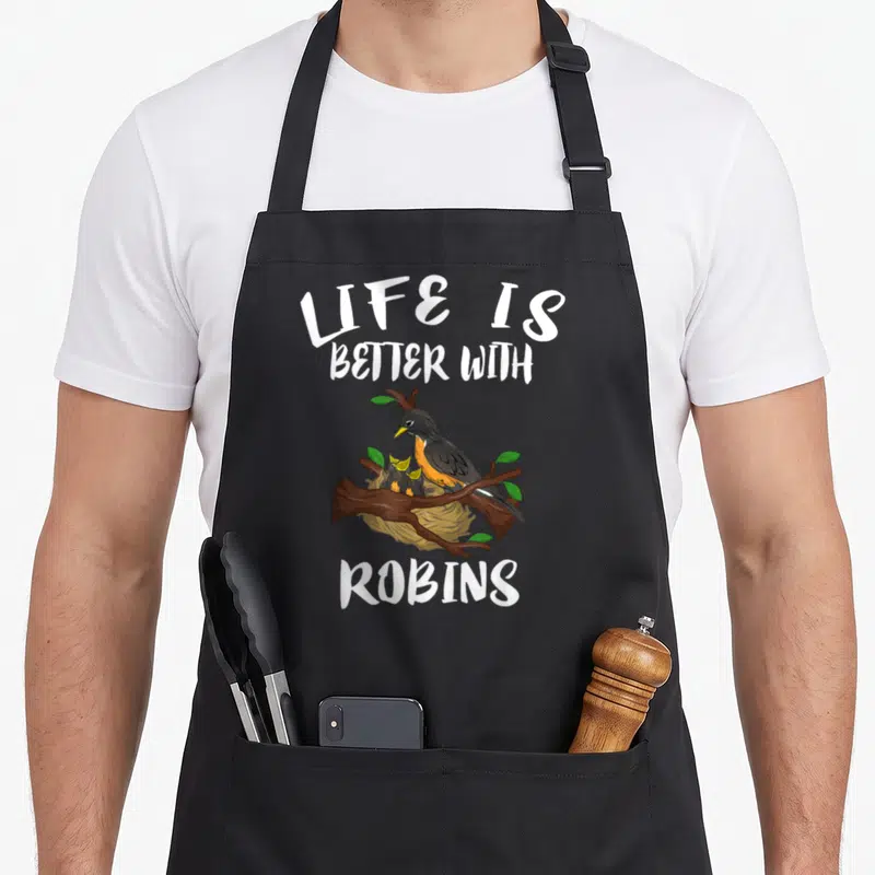 Better bird apron - Cartoon robin perched on a branch above a nest with baby birds and white text reading LIFE IS — shown on a black cotton canvas apron against a white background