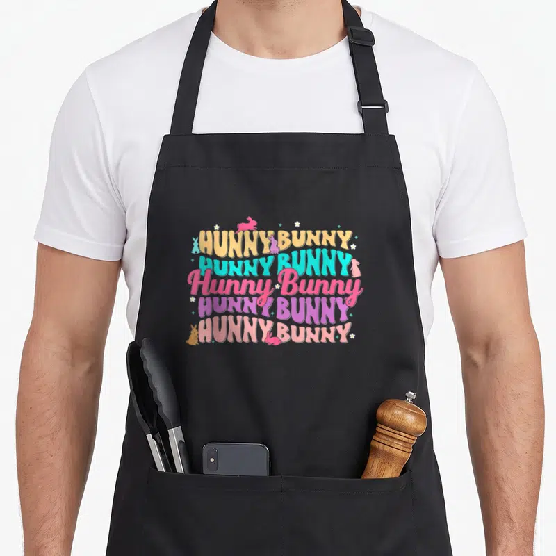 Hunny rabbit bunny apron - Five stacked HUNNY BUNNY text lines in yellow cyan pink purple orange with bunnies — shown on a black cotton canvas apron against a white background