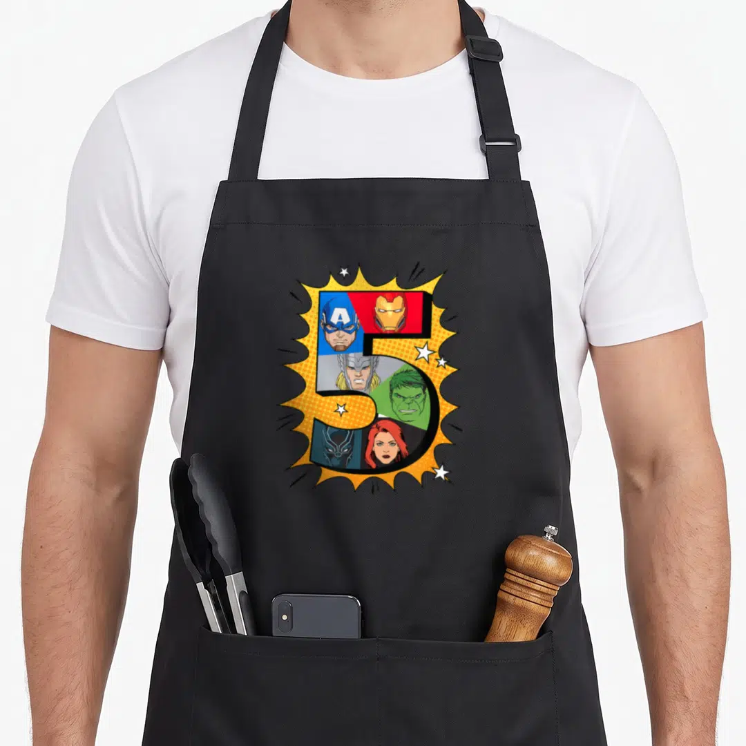 Comic heroe birthday apron - Number 5 filled with six superhero faces inside yellow burst with white stars — shown on a black cotton canvas apron against a white background