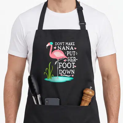 Nana pink flamingo apron - Cartoon flamingo standing in water next to stacked text DON'T MAKE NANA PUT HER FOOT DOWN — shown on a black cotton canvas apron against a white background