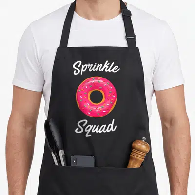 Squad donut sprinkle apron - Pink frosted donut with sprinkles between white Sprinkle Squad script text — shown on a black cotton canvas apron against a white background