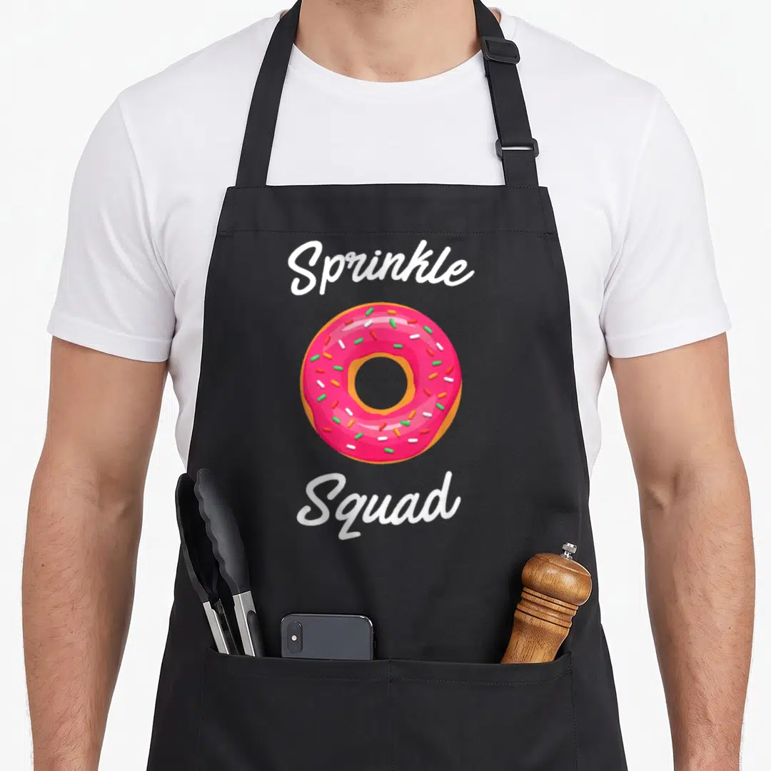 Squad donut sprinkle apron - Pink frosted donut with sprinkles between white Sprinkle Squad script text — shown on a black cotton canvas apron against a white background