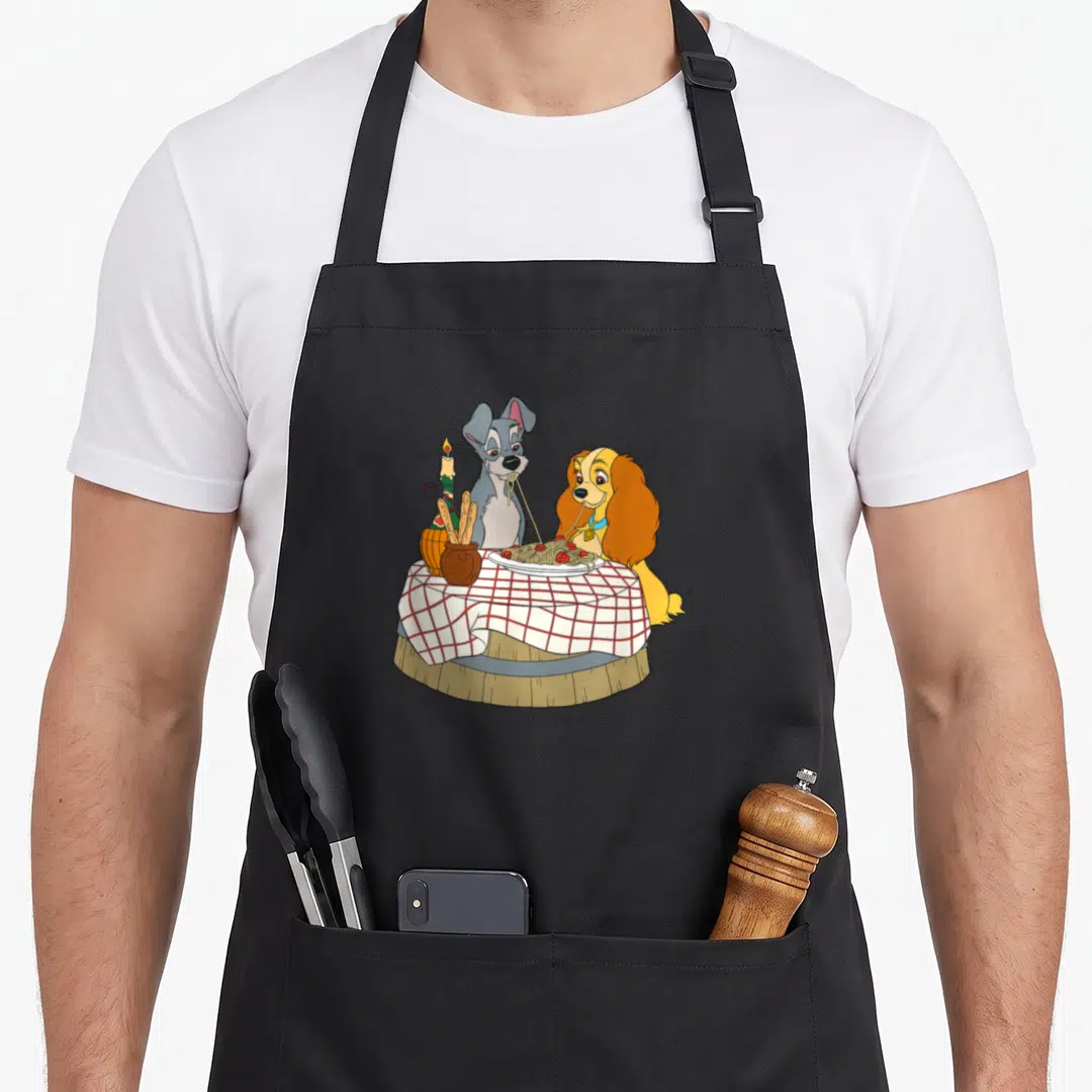 Notte lady and the tramp apron - Two cartoon dogs eating spaghetti at a round table with a candle and breadsticks — shown on a black cotton canvas apron against a white background
