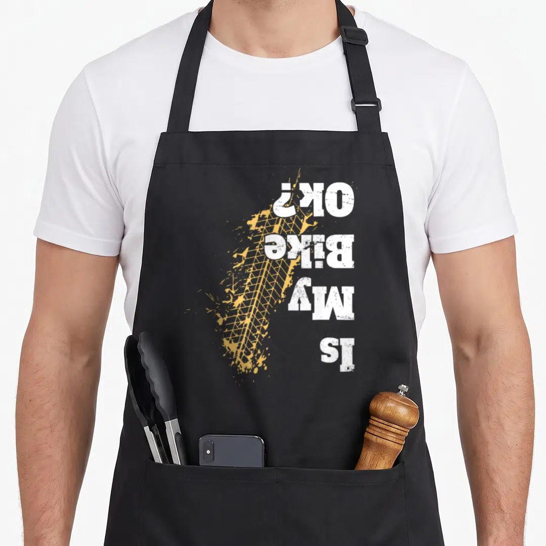 Offroad biker apron - Vertical Is My Bike Ok text over yellow distressed tire track and splatter — shown on a black cotton canvas apron against a white background