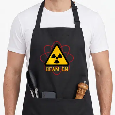 Chemo radiation aprons - Yellow radiation triangle with red orbits and BEAM ON text — shown on a black cotton canvas apron against a white background