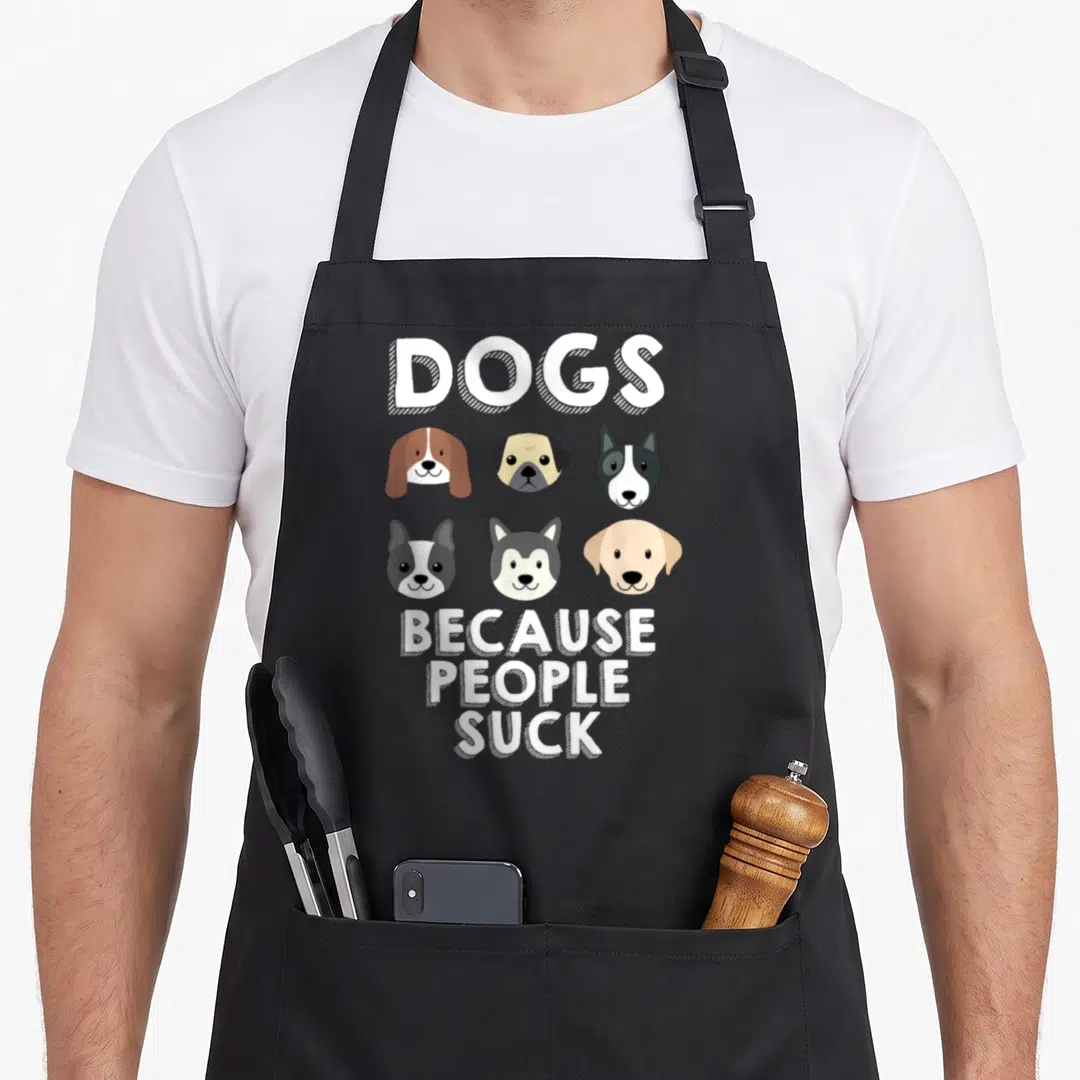 Suck dog apron - Six cartoon dog heads with DOGS BECAUSE PEOPLE SUCK text in white — shown on a black cotton canvas apron against a white background