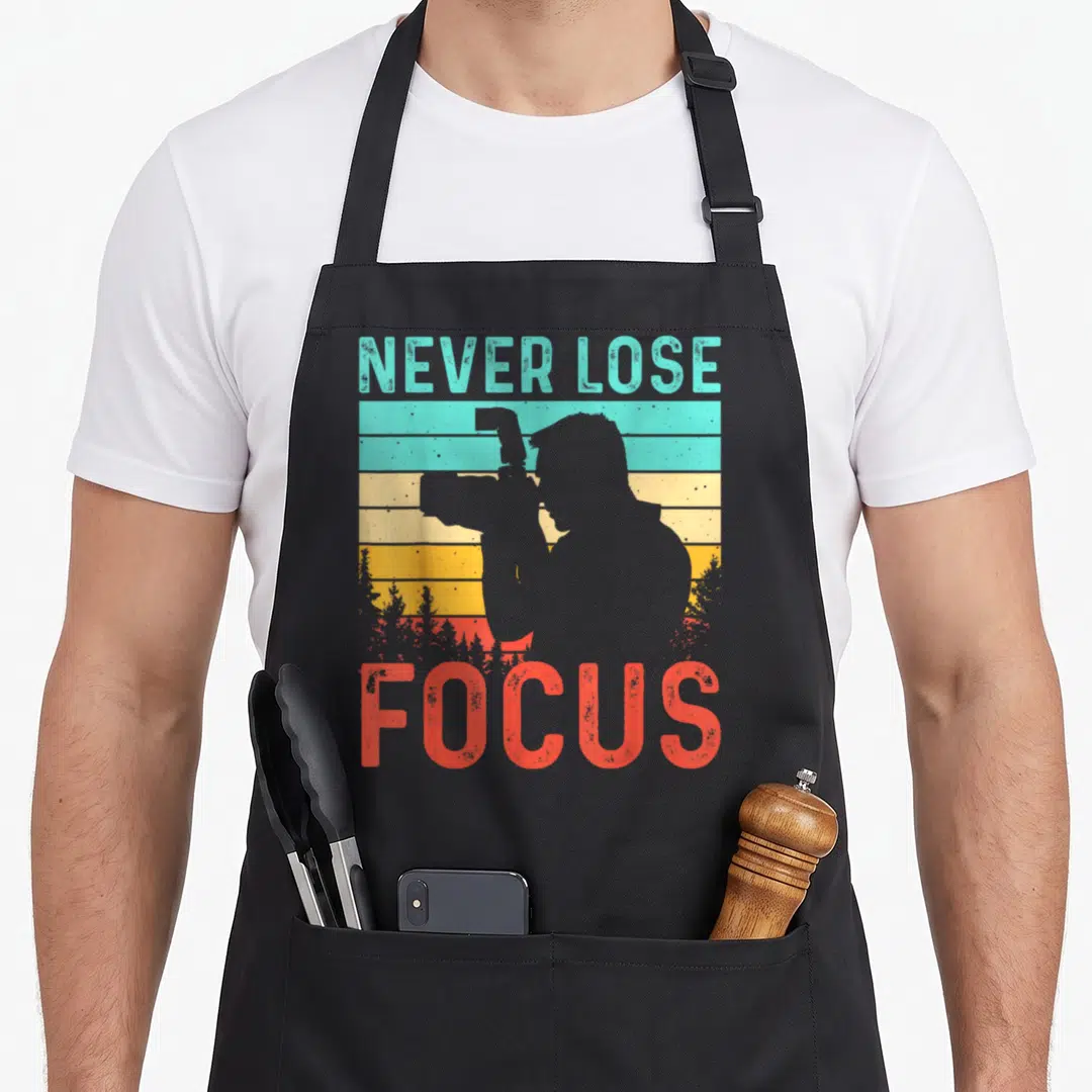 Retro camera photographer apron - NEVER LOSE FOCUS text over striped sunset with photographer silhouette — shown on a black cotton canvas apron against a white background