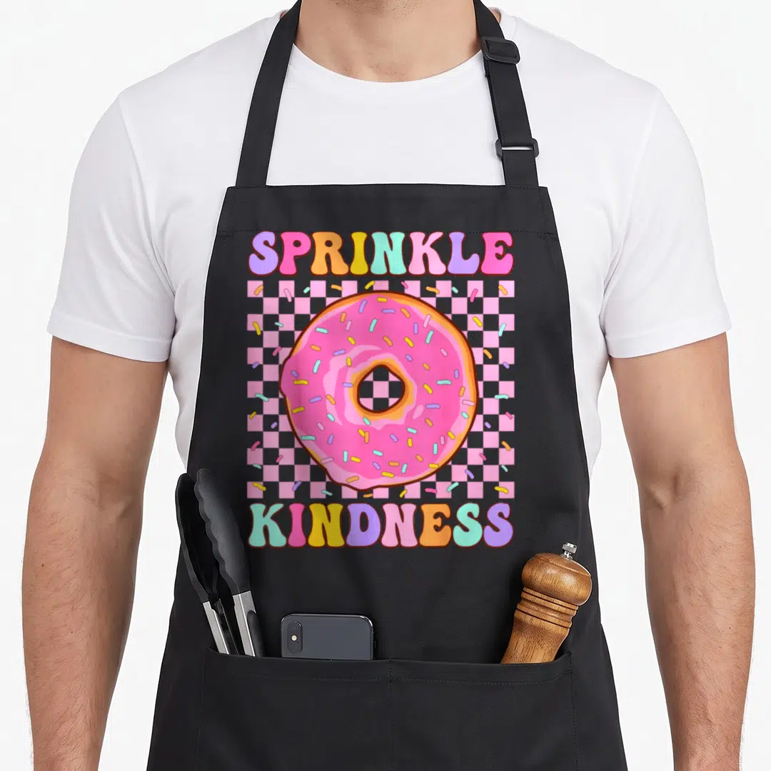 Kindness donut apron - Sprinkle Kindness text surrounding a pink frosted donut on a checkerboard background — shown on a black cotton canvas apron against a white background