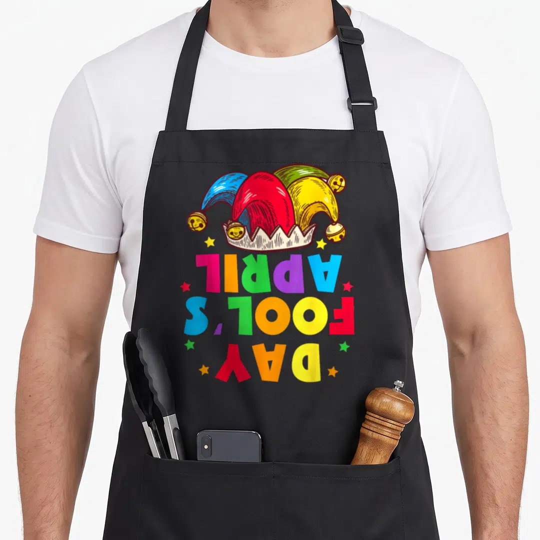 Kit prank apron - Jester hat above vertical APRIL FOOL'S DAY text in red blue yellow green purple orange with stars — shown on a black cotton canvas apron against a white background