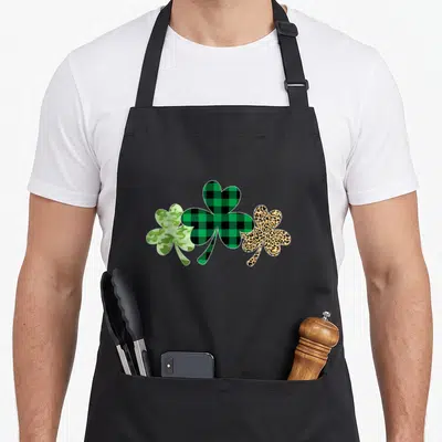 Plaid leopard apron - Three shamrocks in green camo, black plaid, and tan leopard print arranged in a row — shown on a black cotton canvas apron against a white background